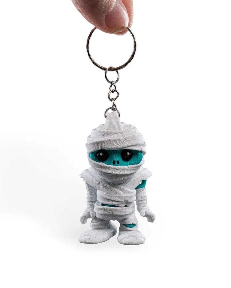 A hand holds a metal ring and chain above a small white wrapped monster figure with a turquoise face.