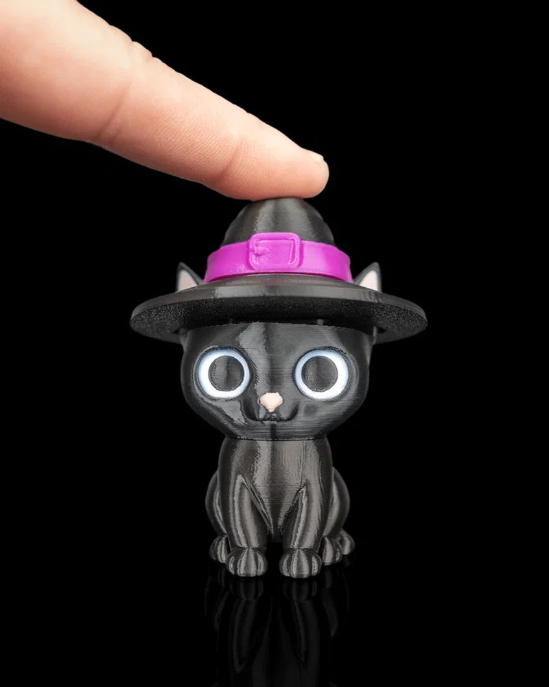 A finger touches the top of a black cat figurine wearing a witch hat with a pink band.